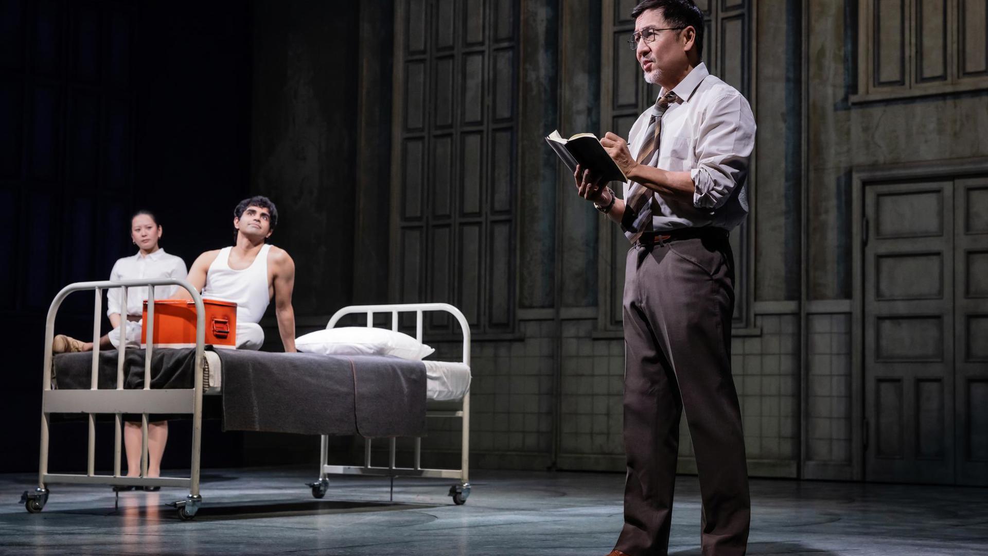 An actor sits on a bed set piece. An actor dressed as a nurse stands in the background. An actor portraying a doctor stands in the foreground writing in a journal.