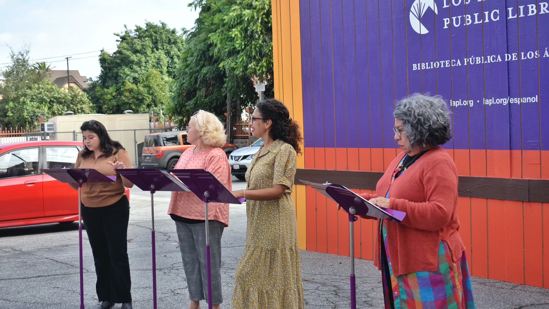 A group of actors stand outside in front of music stands reading through a play. 