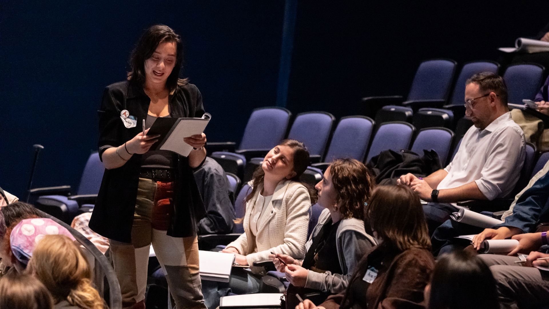 A person stands reading a script with a folks listening in theatre seats. 