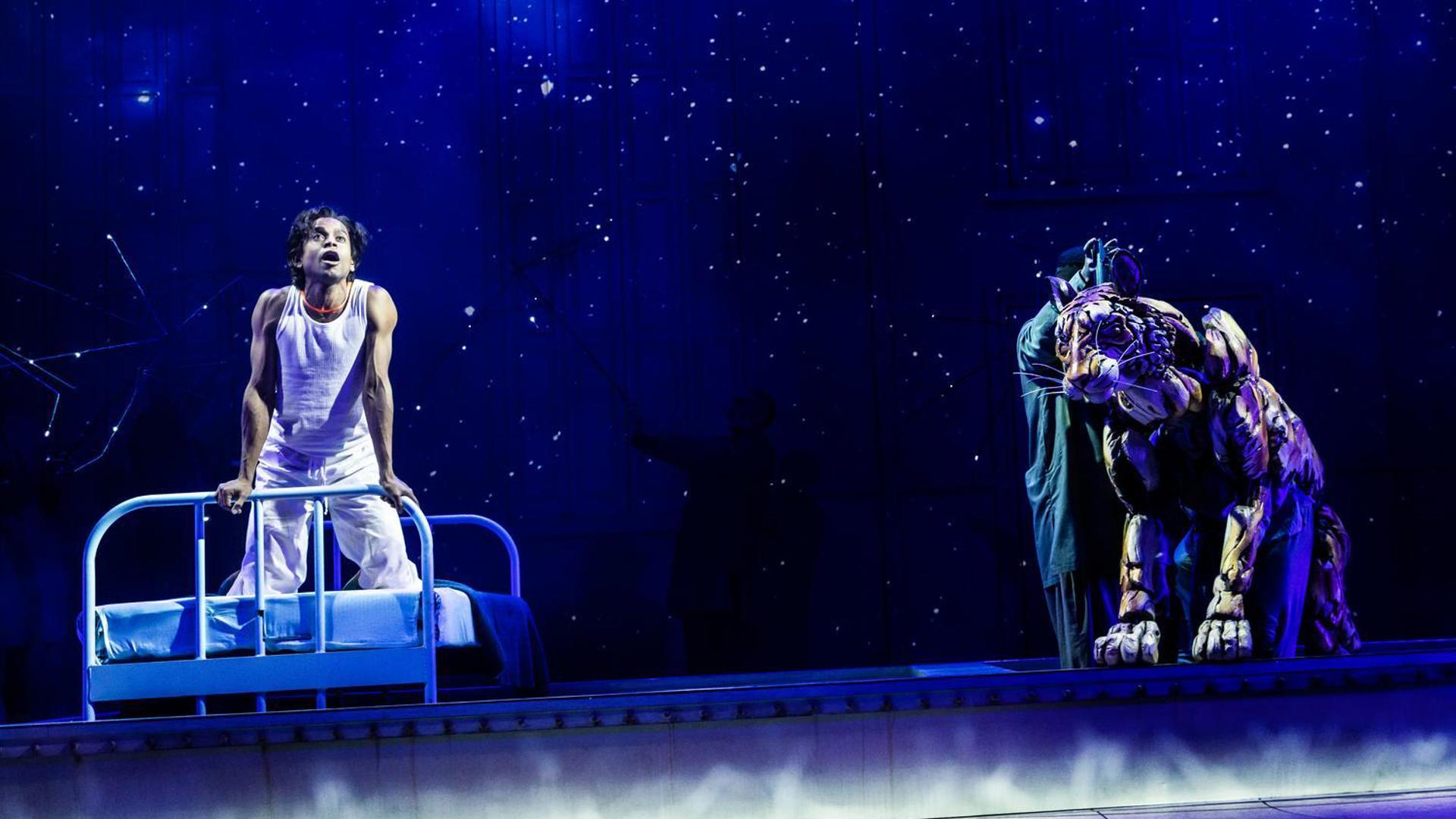 Actor on a hospital bed looks up in awe under a starry night sky, while a tiger puppet, manipulated by performers, sits nearby in a scene from 'Life of Pi.