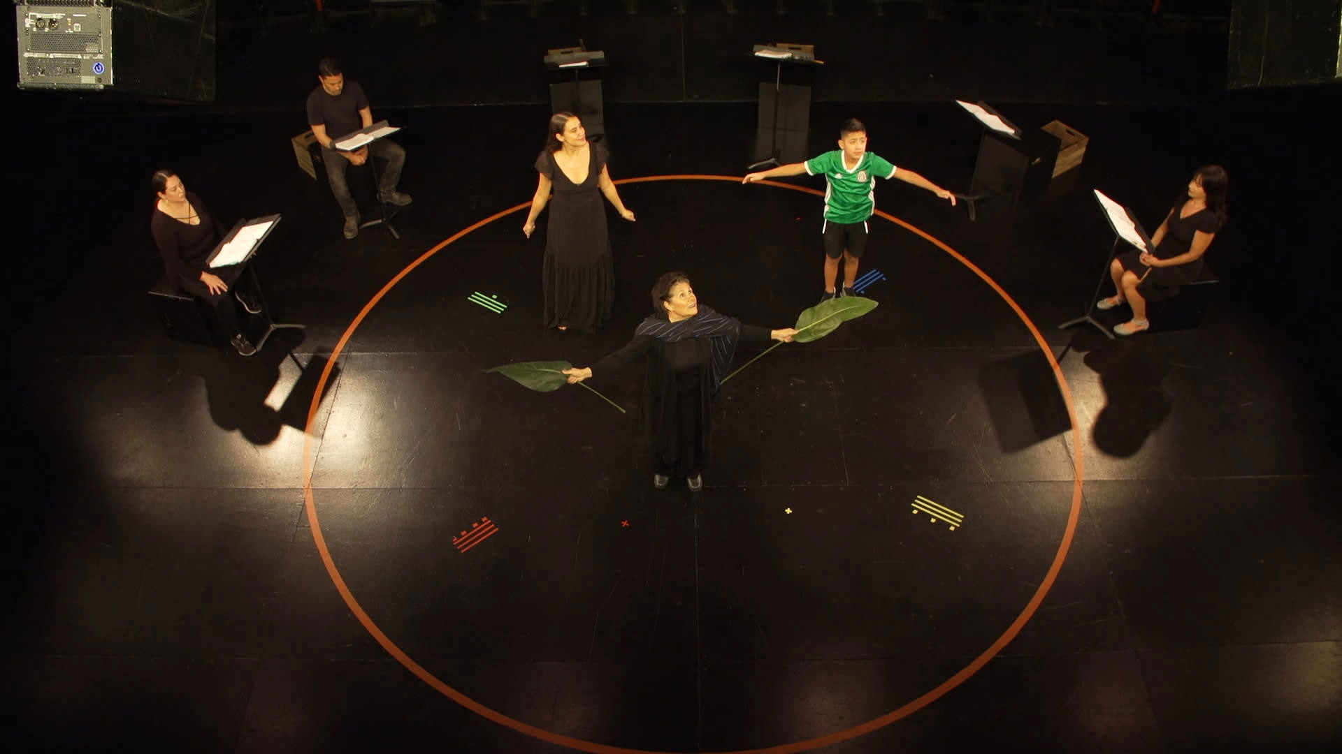 Actors rehearse a play inside the rehearsal room. Three actors stand within a circle with their arms out, while three other actors sit outside the circle.