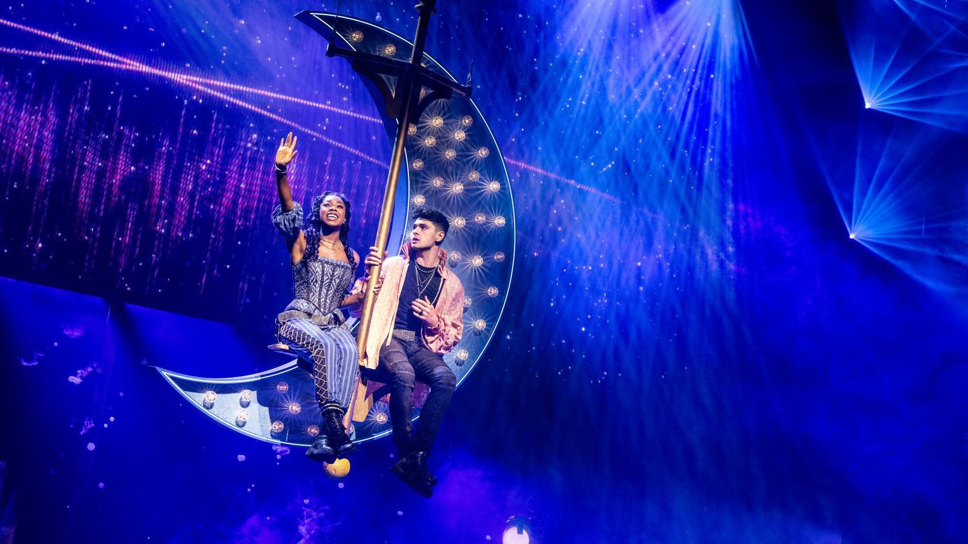 Rachel Simone Webb and Michael Canu sit in a lit-up crescent moon that is suspended from the ceiling.