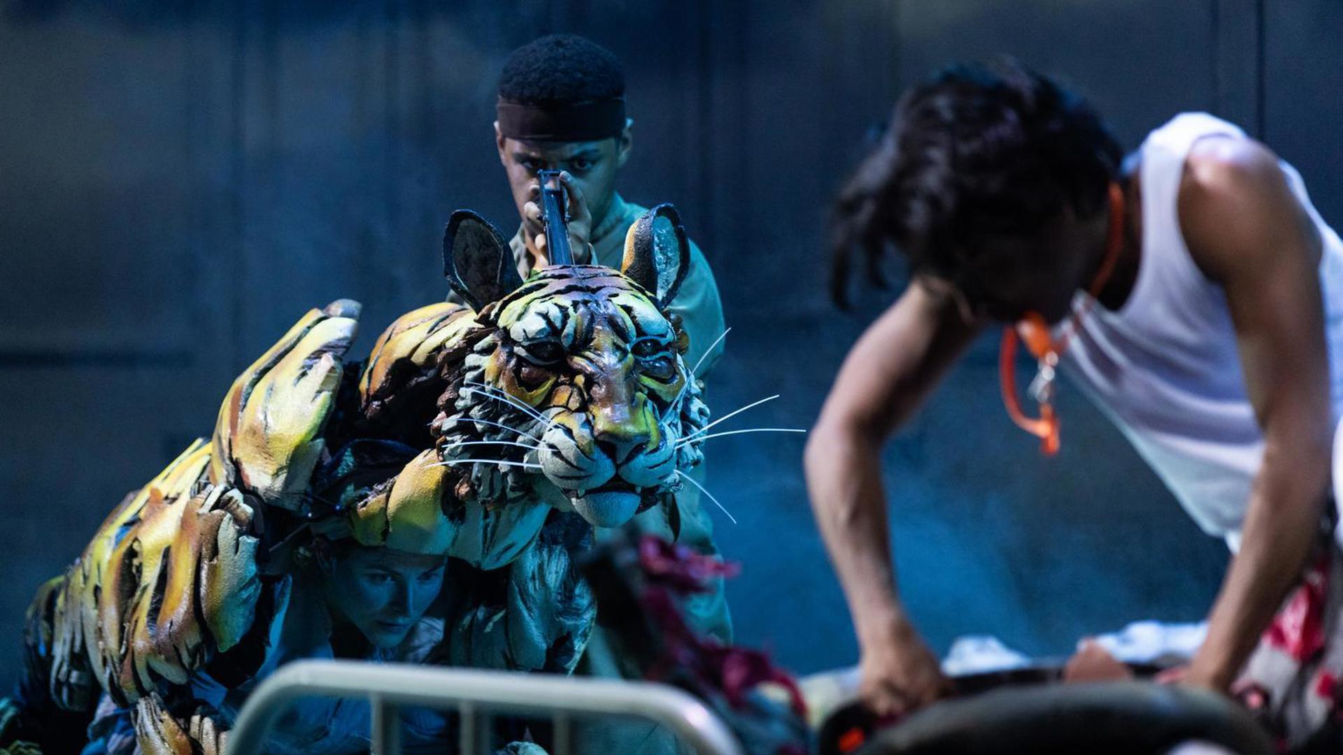 Actors manipulate a realistic tiger puppet on stage during a dramatic scene from 'Life of Pi,' with atmospheric lighting and intense expressions