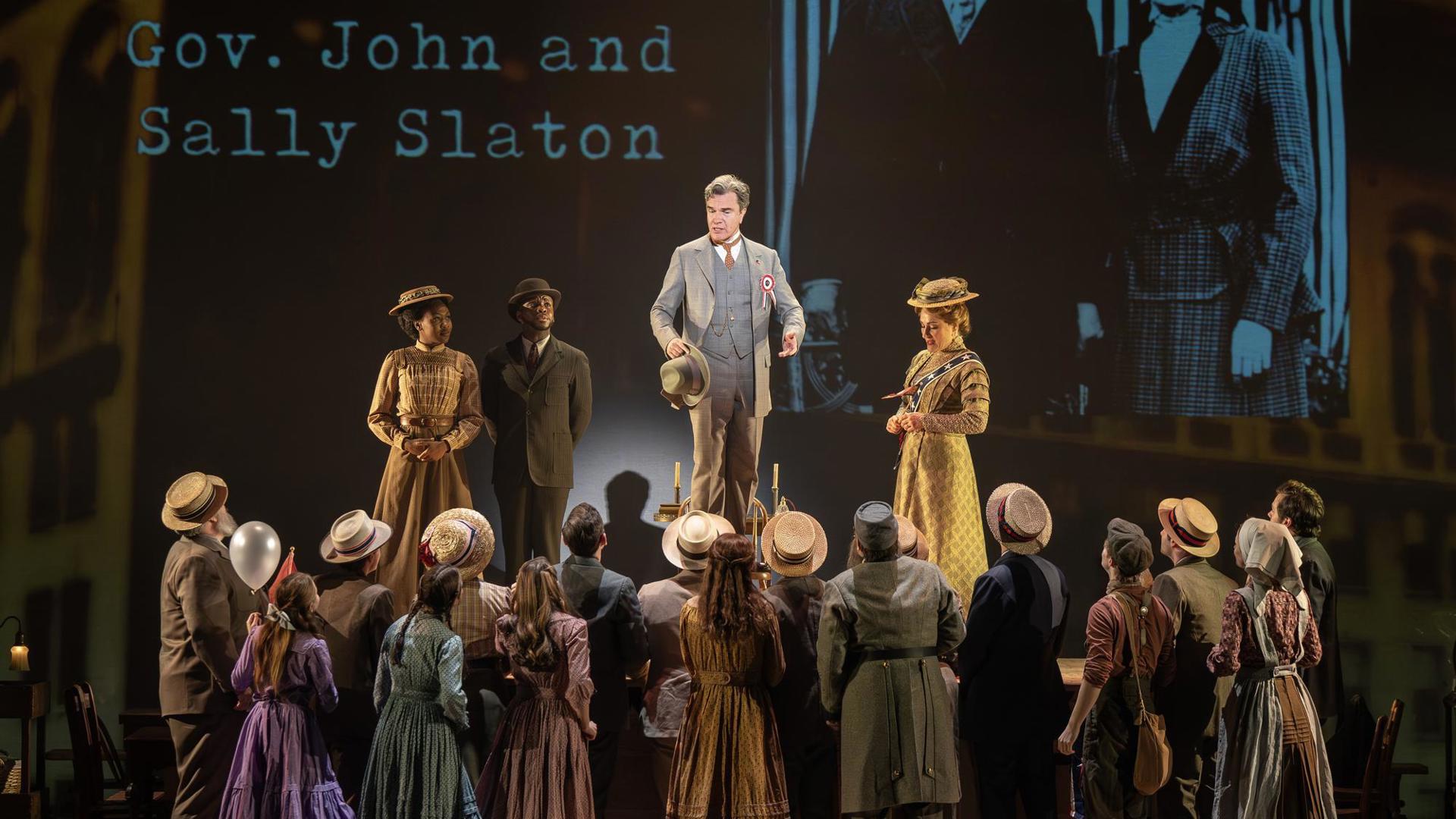 A man on a platform speaks to a large crowd. In the background, a projection that reads "Gov. John and Sally Slaton."