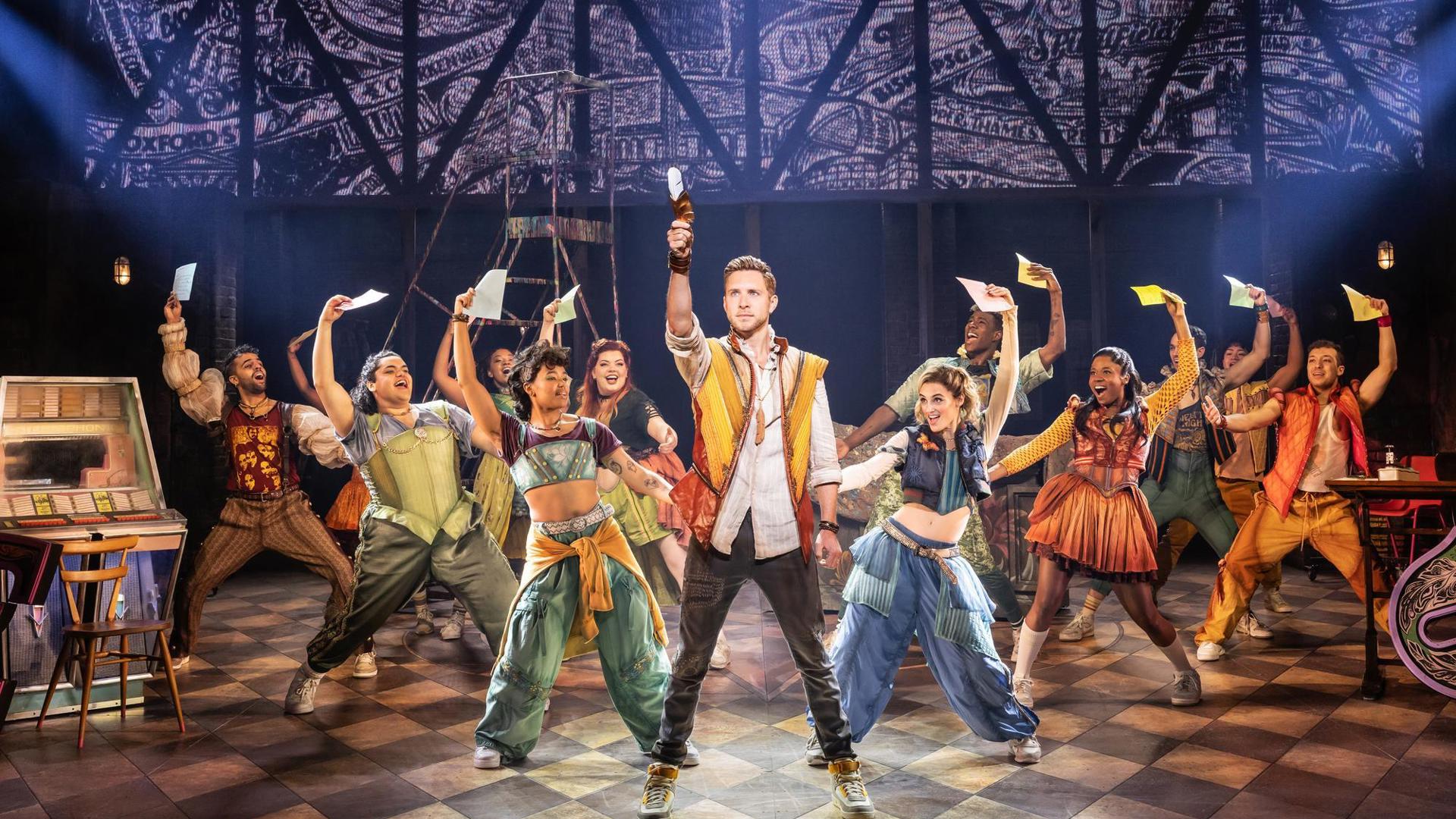 Corey Mach stands center stage surrounded by dancers. They all lift multicolored papers over their heads.