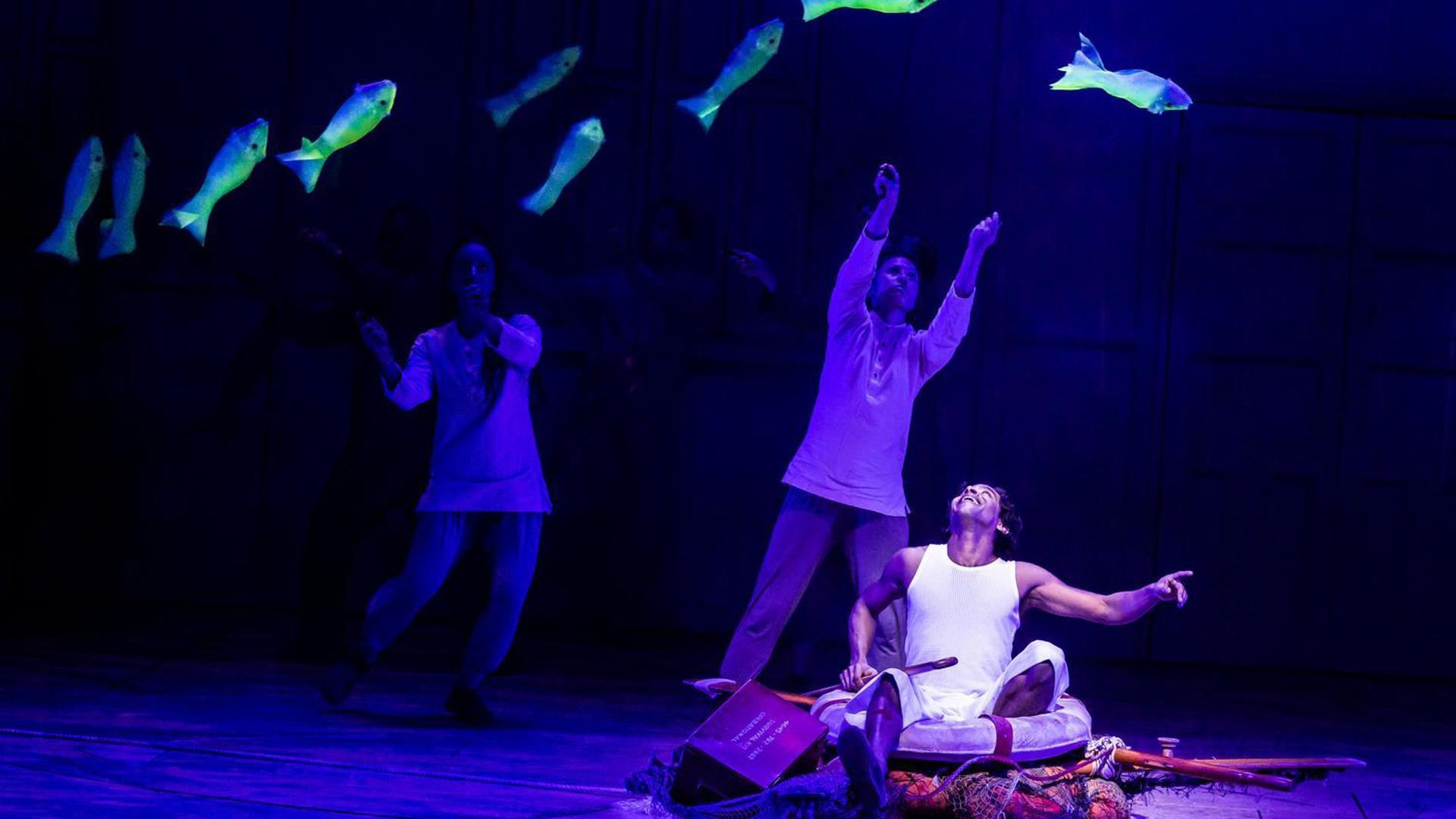 ctors perform under blue lighting, with glowing fish suspended in the air, as a seated character gestures upward in a scene from 'Life of Pi.'