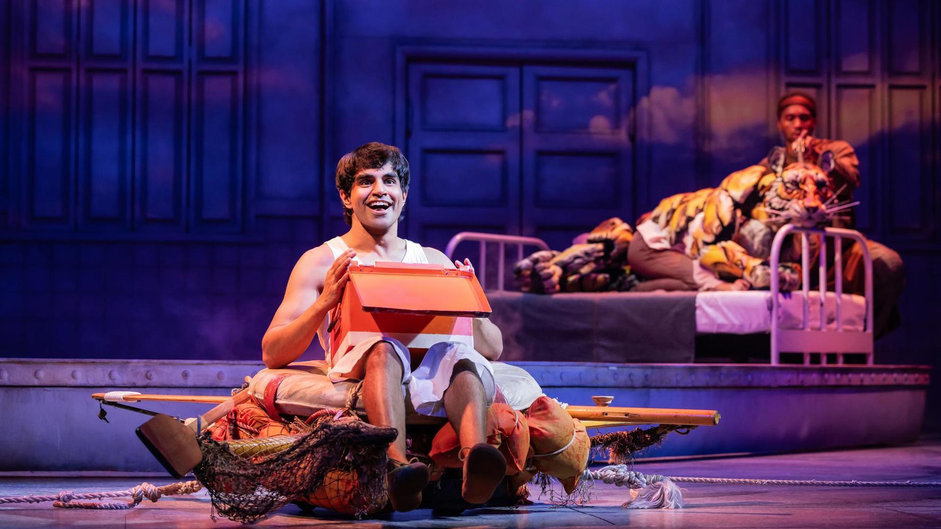 An actor sits on the stage holding an open box. In the background, a puppeteer operates a tiger puppet on a bed set piece.