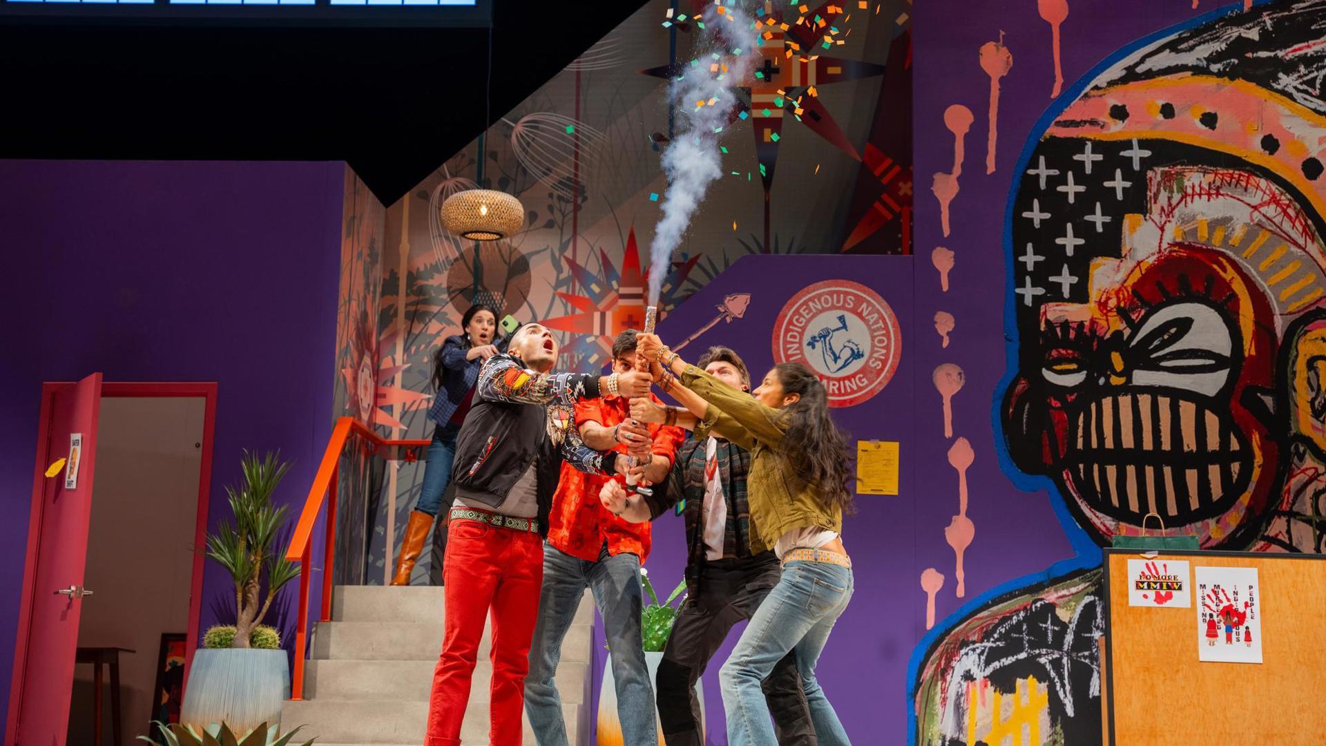 4 people stand on a colorful stage adorned with vibrant Indigenous-inspired artwork, popping confetti, one person stands in the back capturing the moment on a phone.