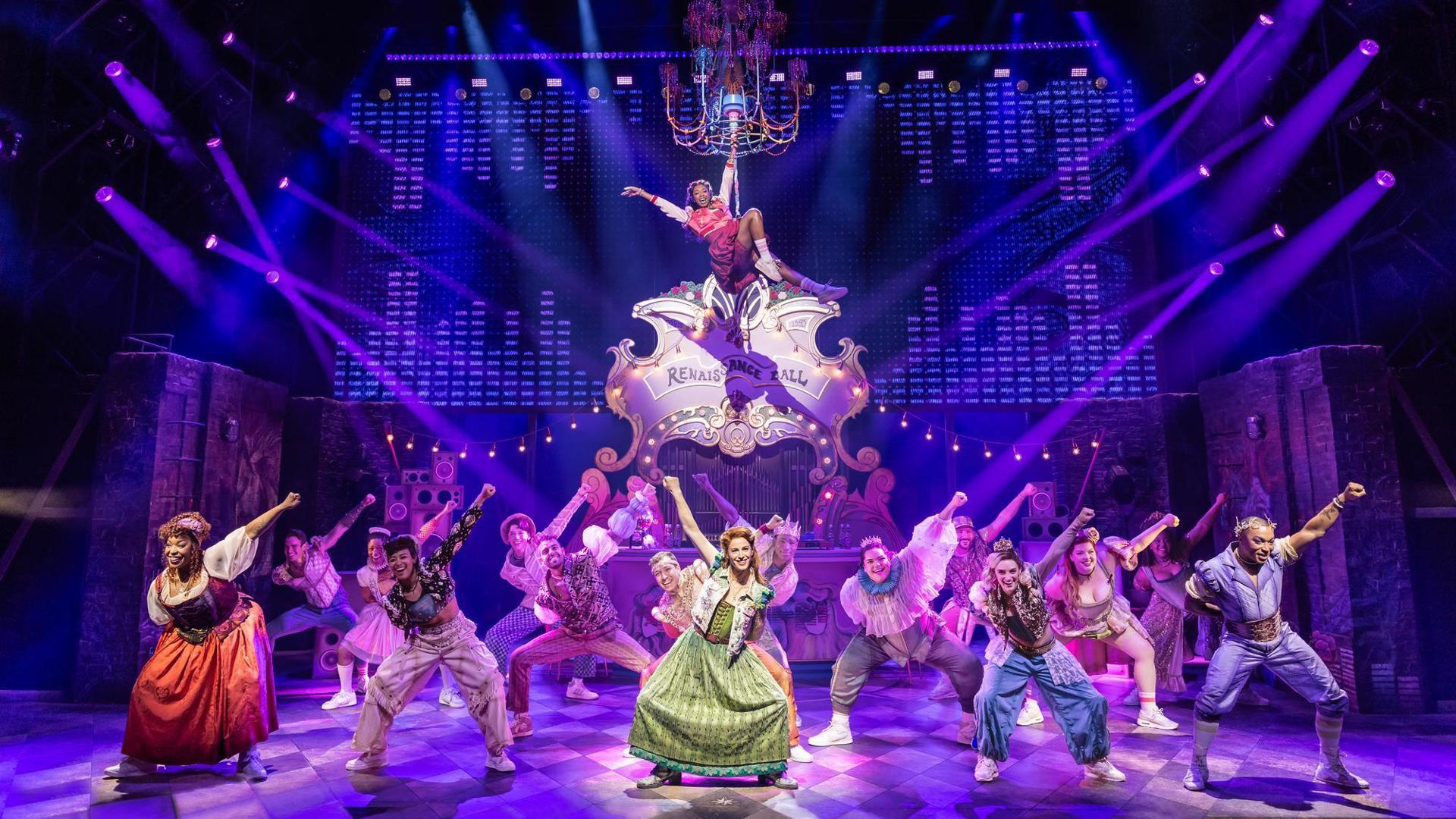 Company of &Juliet dance with one arm up over their heads. Rachel Simone Webb hangs from a chandelier center stage.