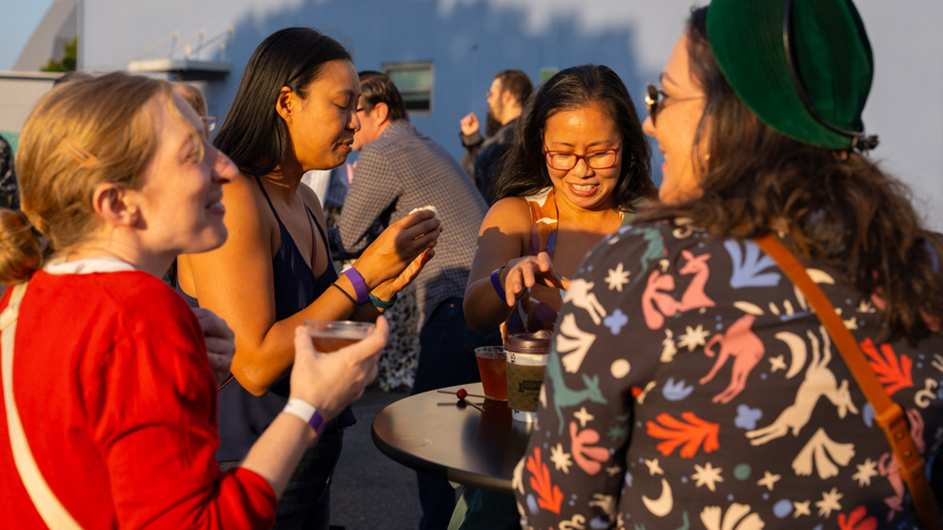 A group a people socialize with drinks in their hands around a high-top table. 