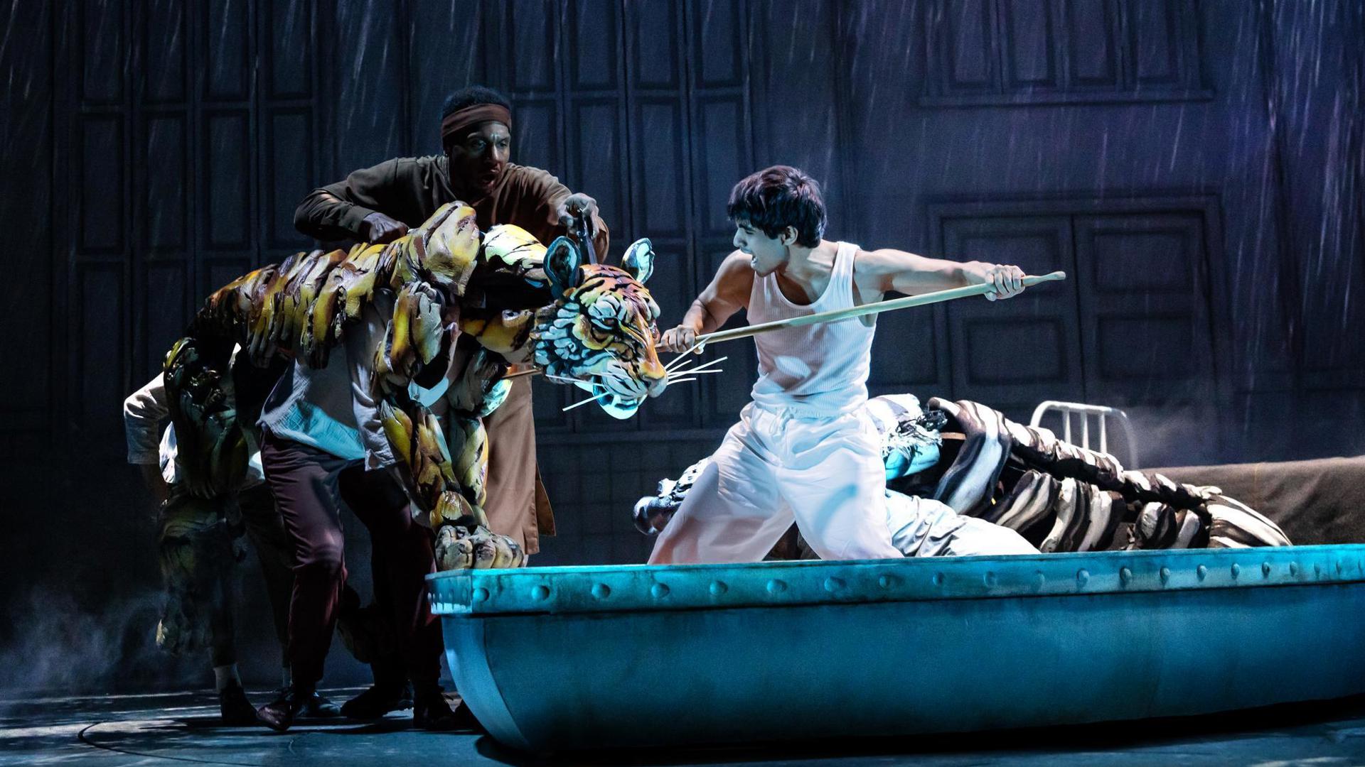 A puppeteer operates a tiger puppet being fought off by an actor, standing in a lifeboat, holding an oar.