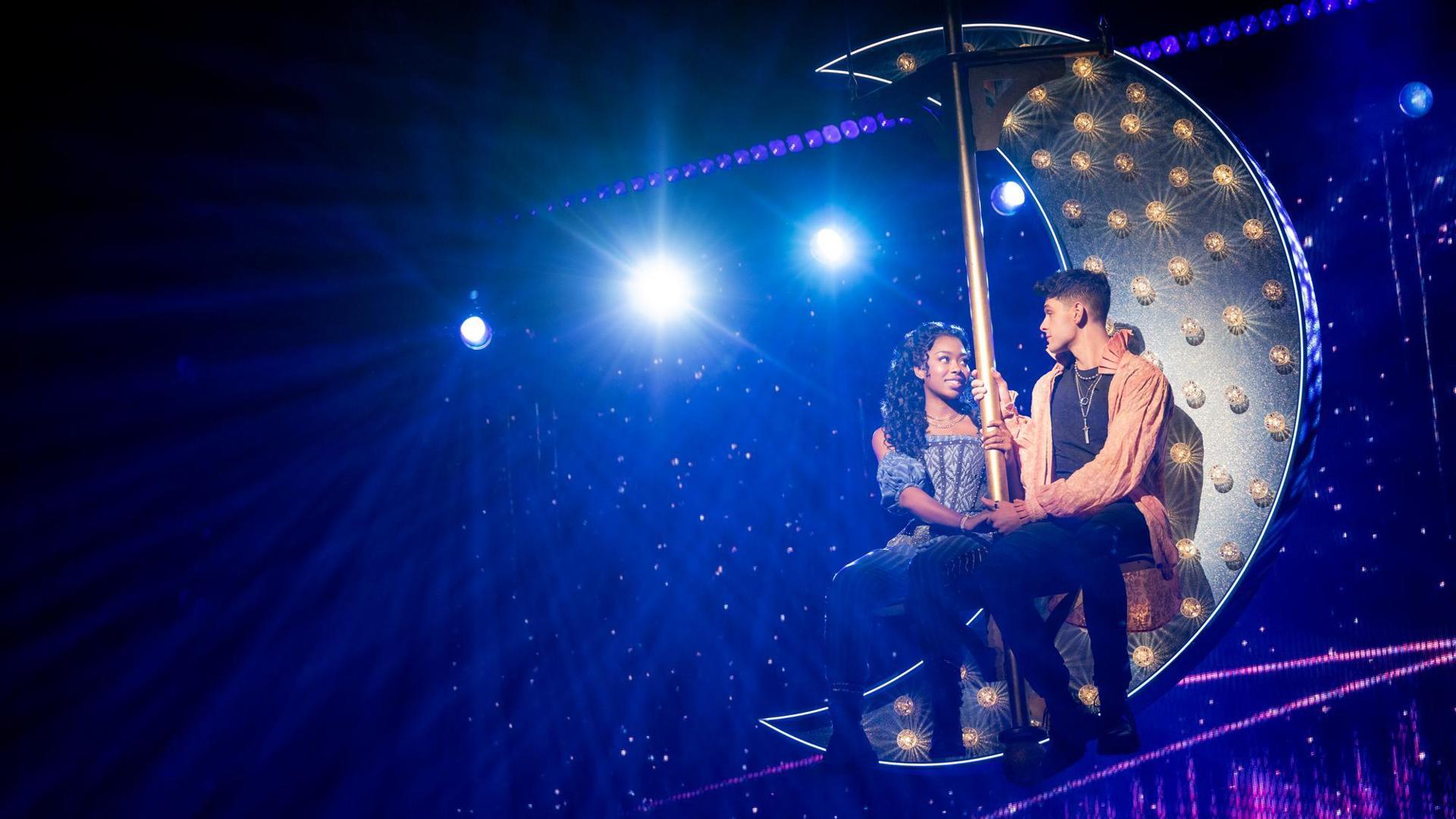 Rachel Simone Webb and Michael Cann sit in a light-up crescent moon that  is being suspended from the ceiling.