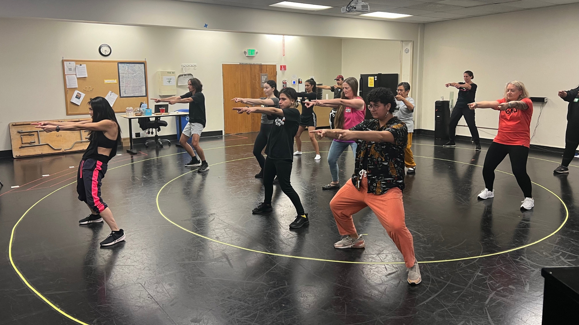 A room of people participate in a workshop, moving with their arms pointing straight forward. 