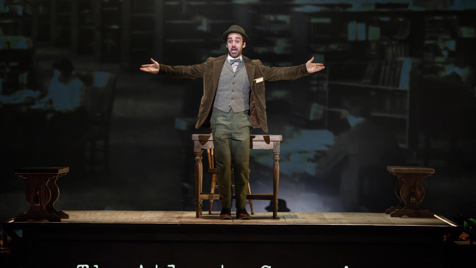 A man in 1910s attire speaking to the audience with his arms out. He stands on a platform that says "The Atlanta Georgian Newspaper Offices."