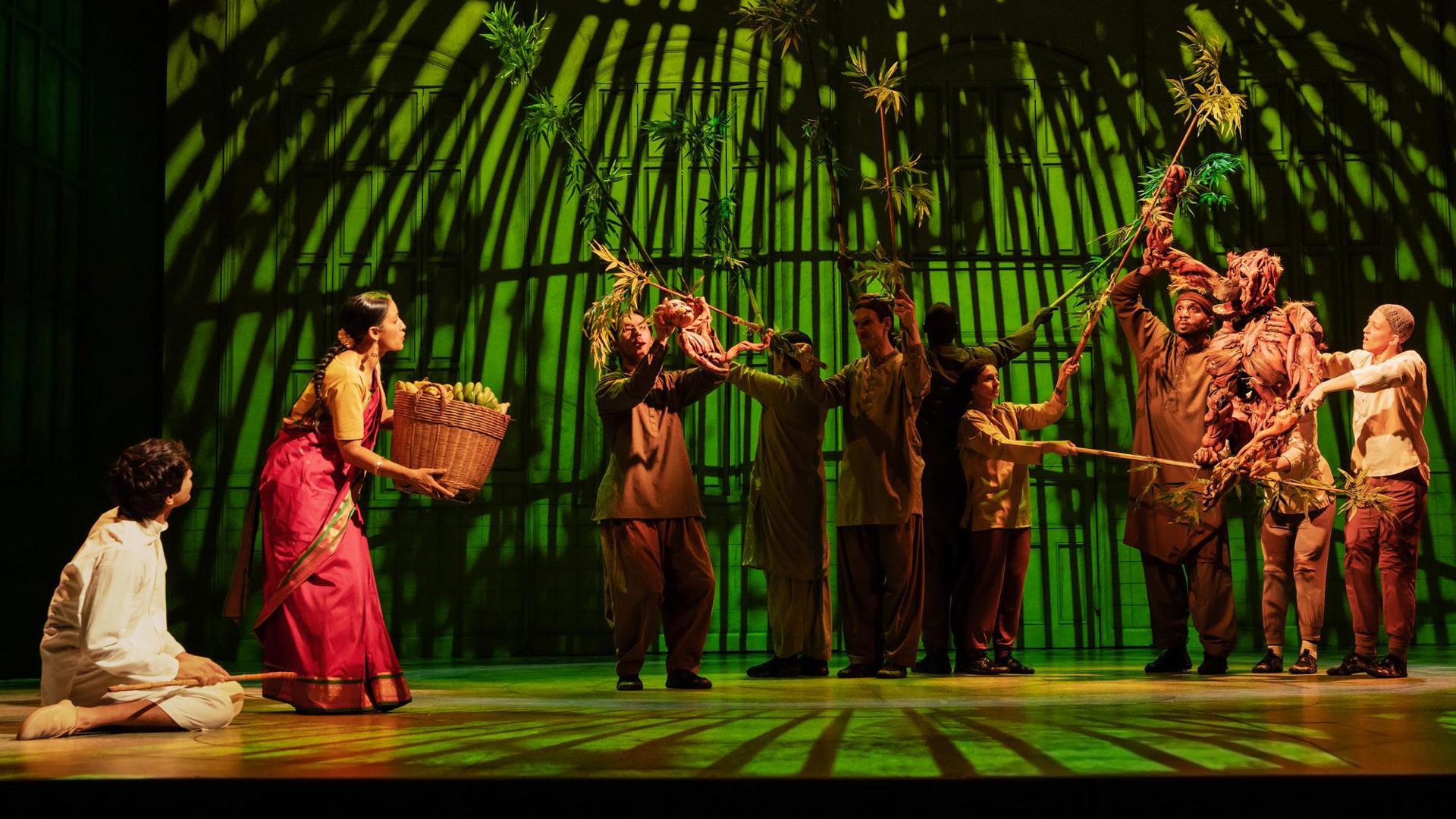 An actor sits on their knees while another actor stands, holding a basket of fruit. Ensemble members hold tree branches, emulating a tree.