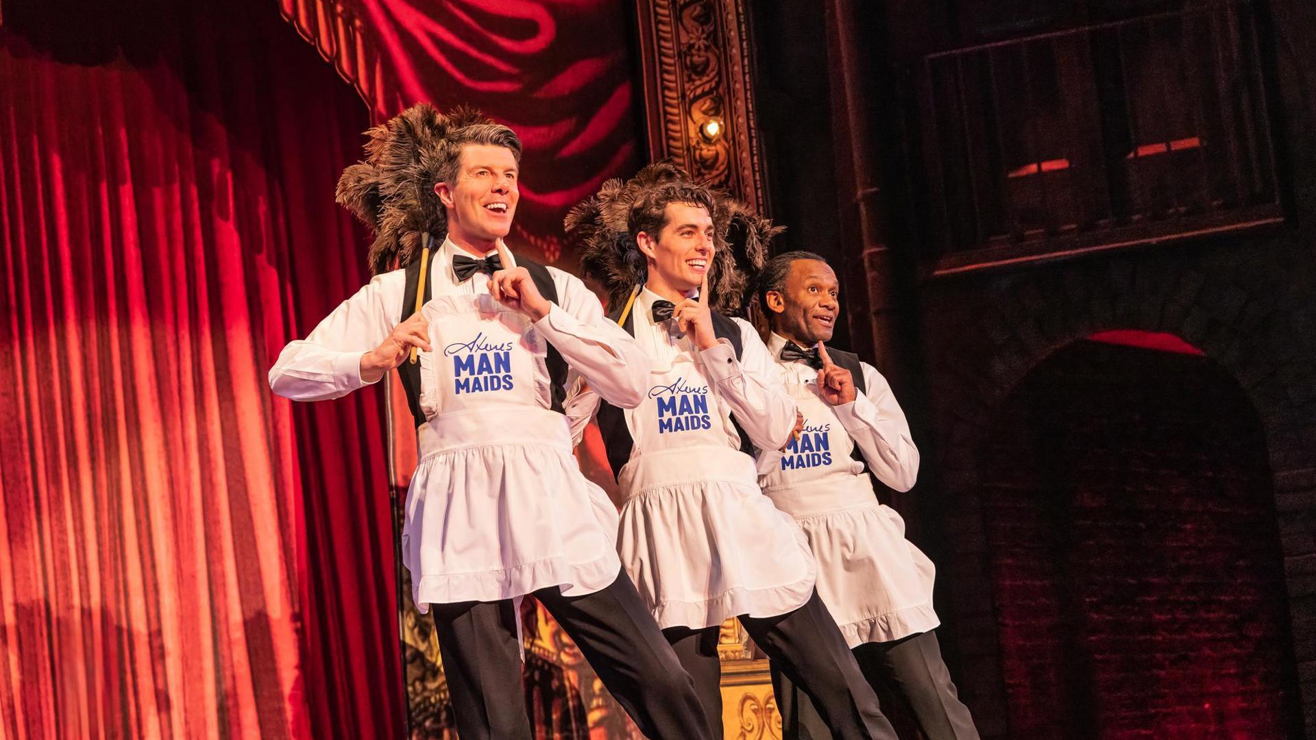 Three performers wearing aprons that read "Steve's Man Maids" hold feather dusters over their shoulders and hold a finger to their lips.