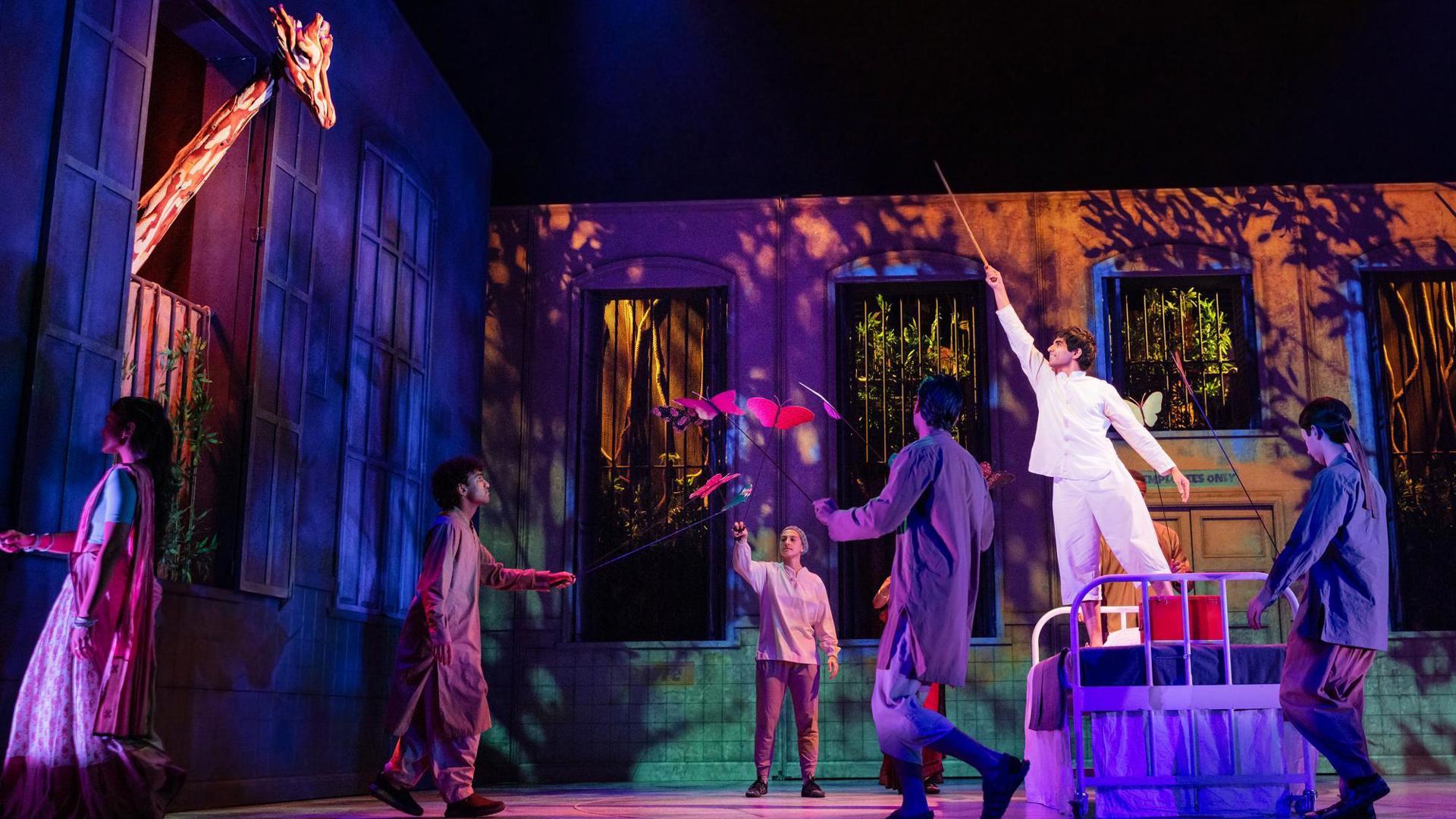 Ensemble members walk around the stage. Some hold butterfly puppets. An actor stands on a bed set piece pointing a stick toward a giraffe puppet that pokes its head out of a window.