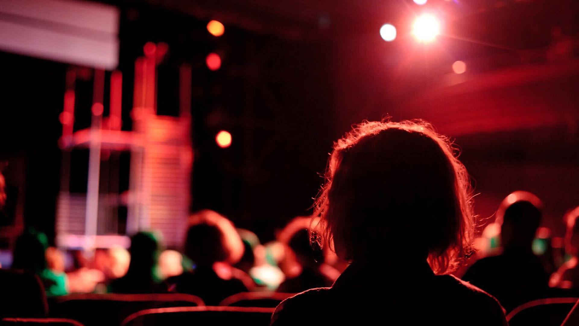 The silhouette of an individual seated in a theatre illuminated by a red light.