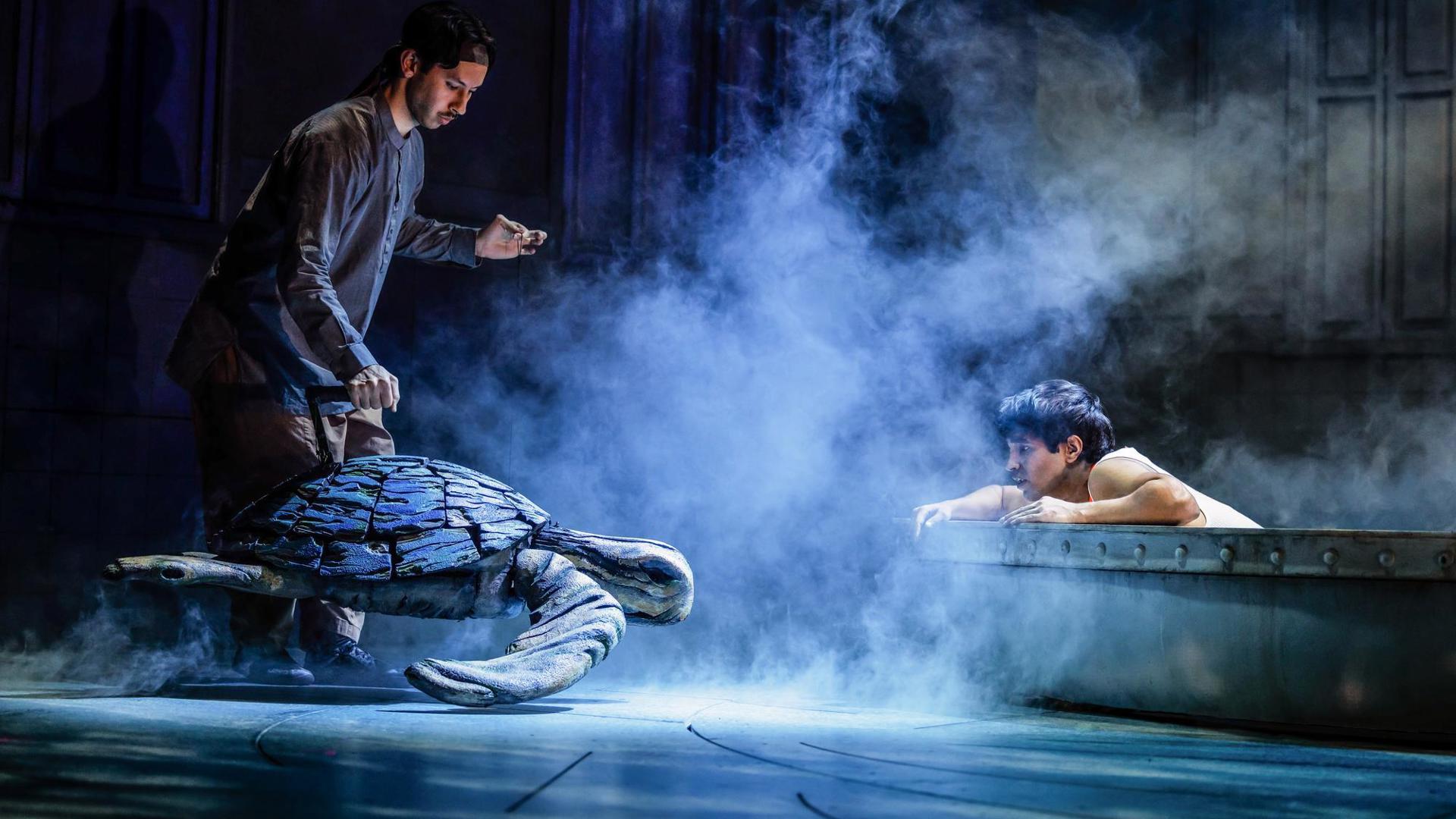 An actor kneels in a lifeboat looking toward a turtle puppet being operated by a puppeteer.