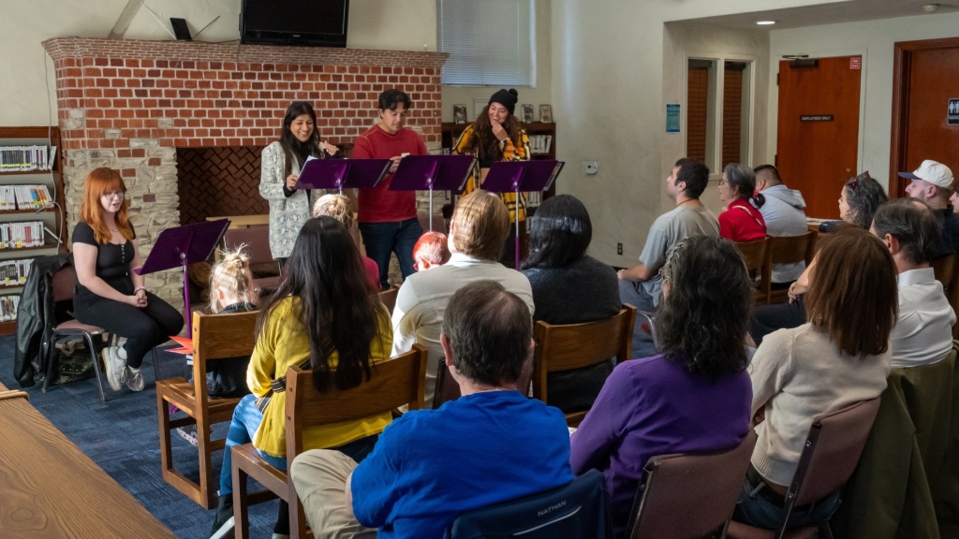 3 actors stand  and 1 sits infront of purple music stands performing to an audience. 