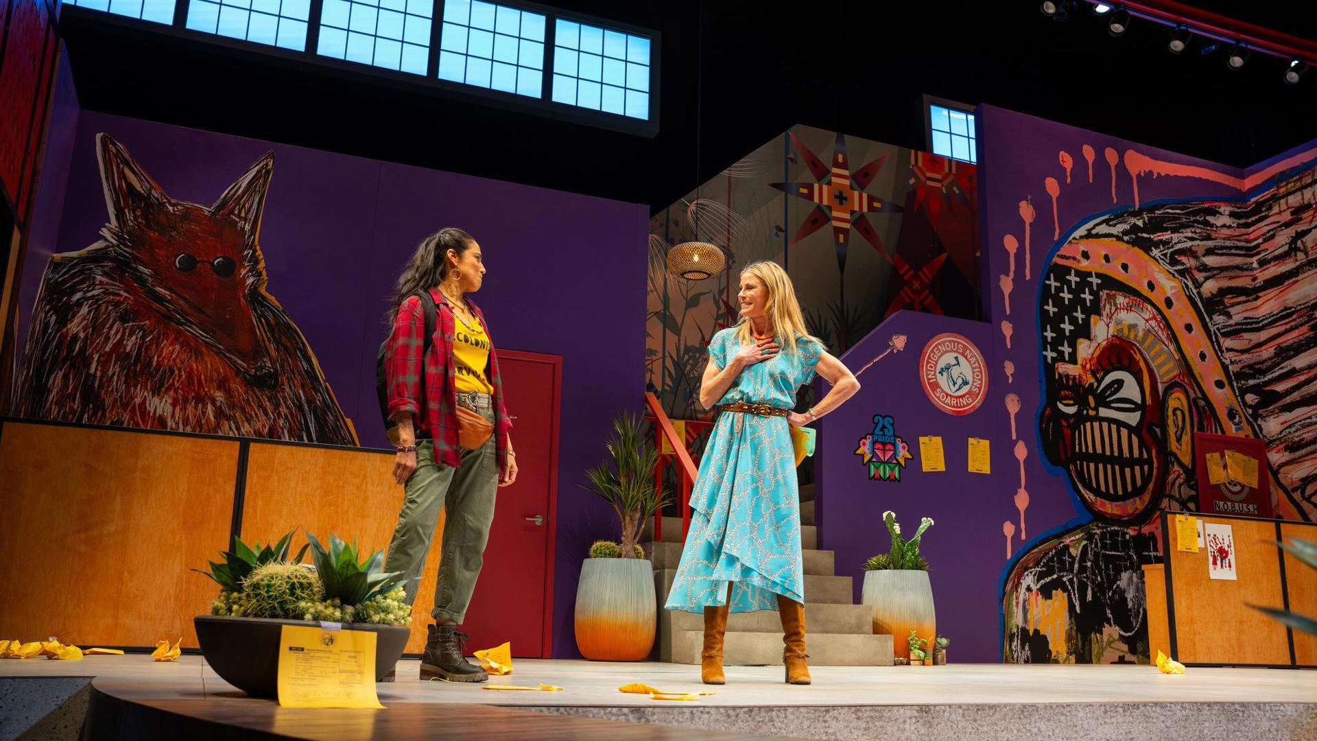 Two people engaged in conversation stand on a colorful stage adorned with vibrant Indigenous-inspired artwork.