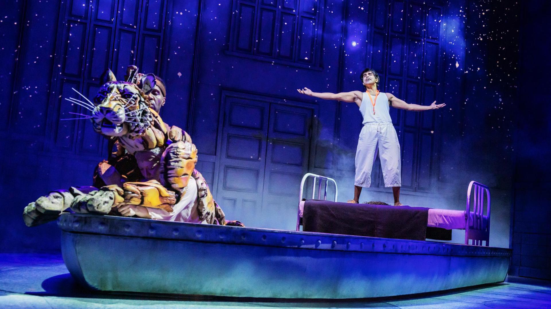 In the background, an actor stands on a bed set piece with their arms out. In the foreground, a puppeteer sits in a lifeboat set piece operating a tiger puppet.