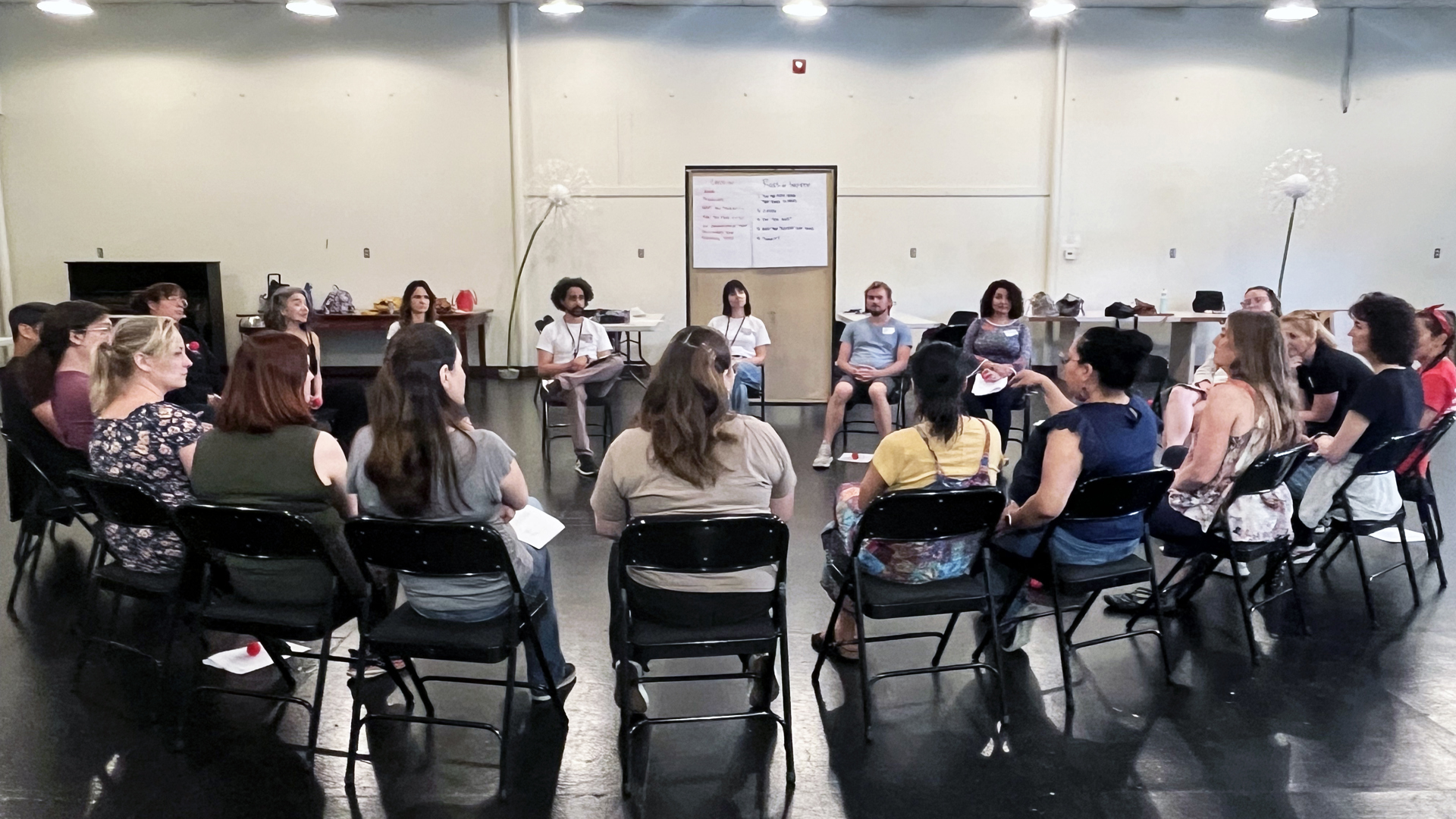 Several educators sit in circle in a rehearsal space actively engaging with each other.  