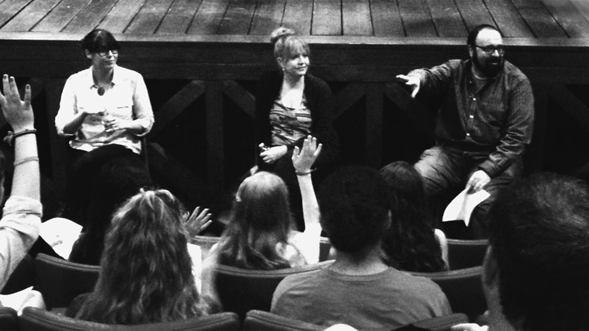 theatre interior, the audience raises their hands to asked questions to a panel of three positioned in front of the stage. 