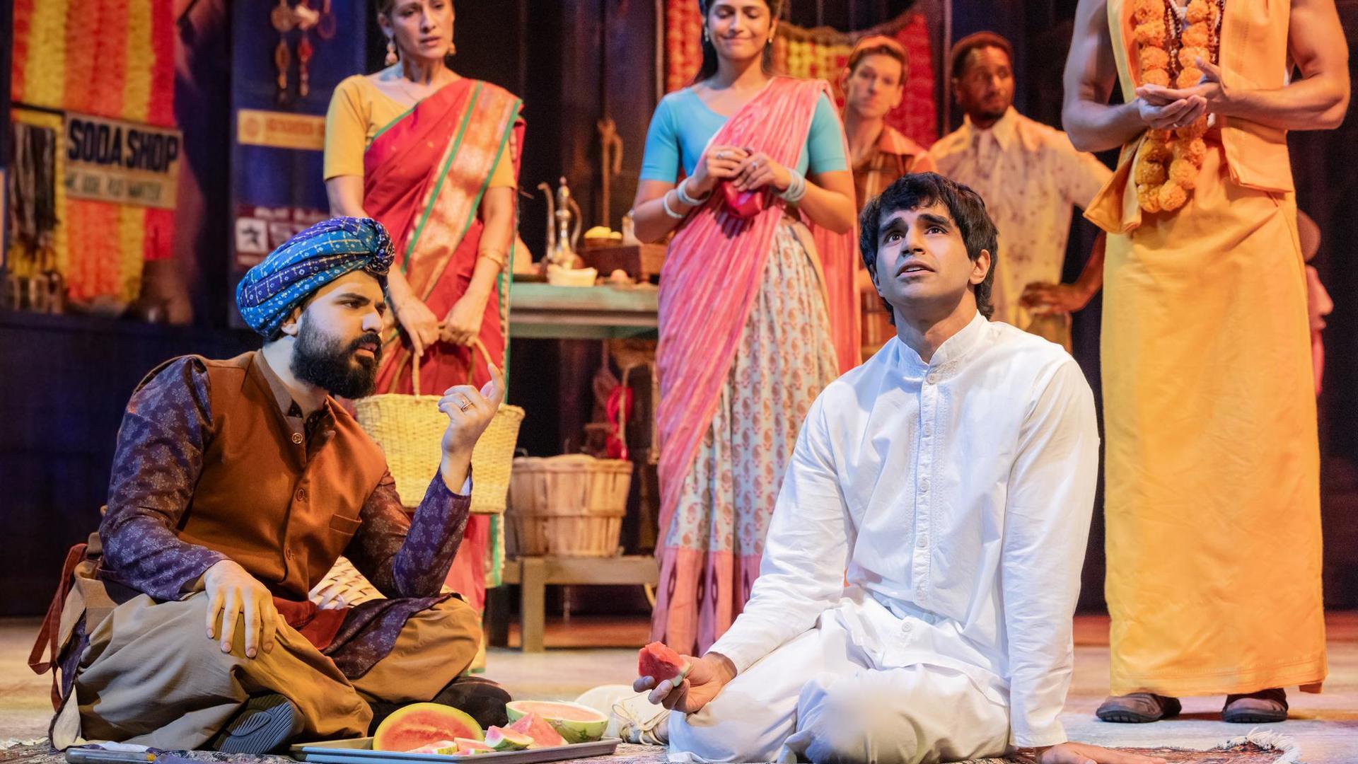 Two actors sit downstage on a rug, eating watermelon. Five ensemble members watch them in the background.