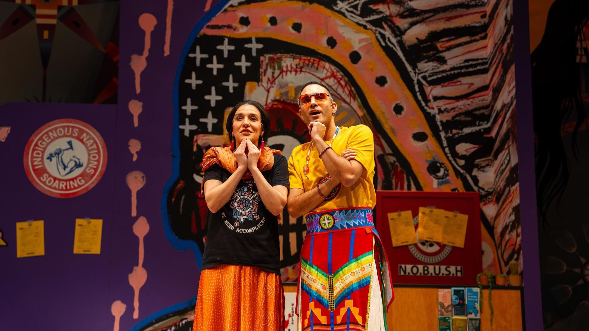 Two people look into the distance stand on a colorful stage adorned with vibrant Indigenous-inspired artwork.