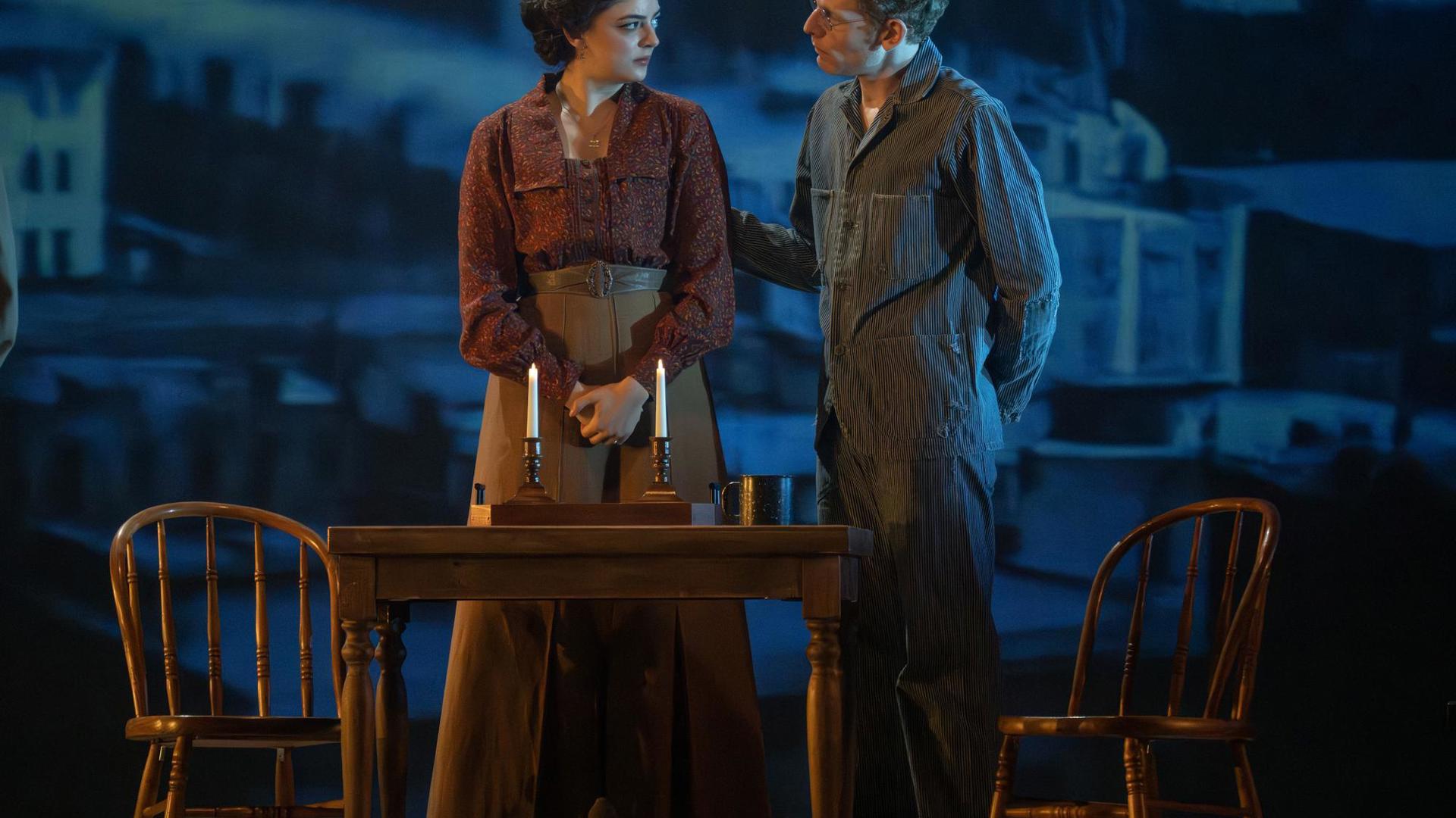 A man and woman standing at a dining table with candles on it. His hand is on her back.