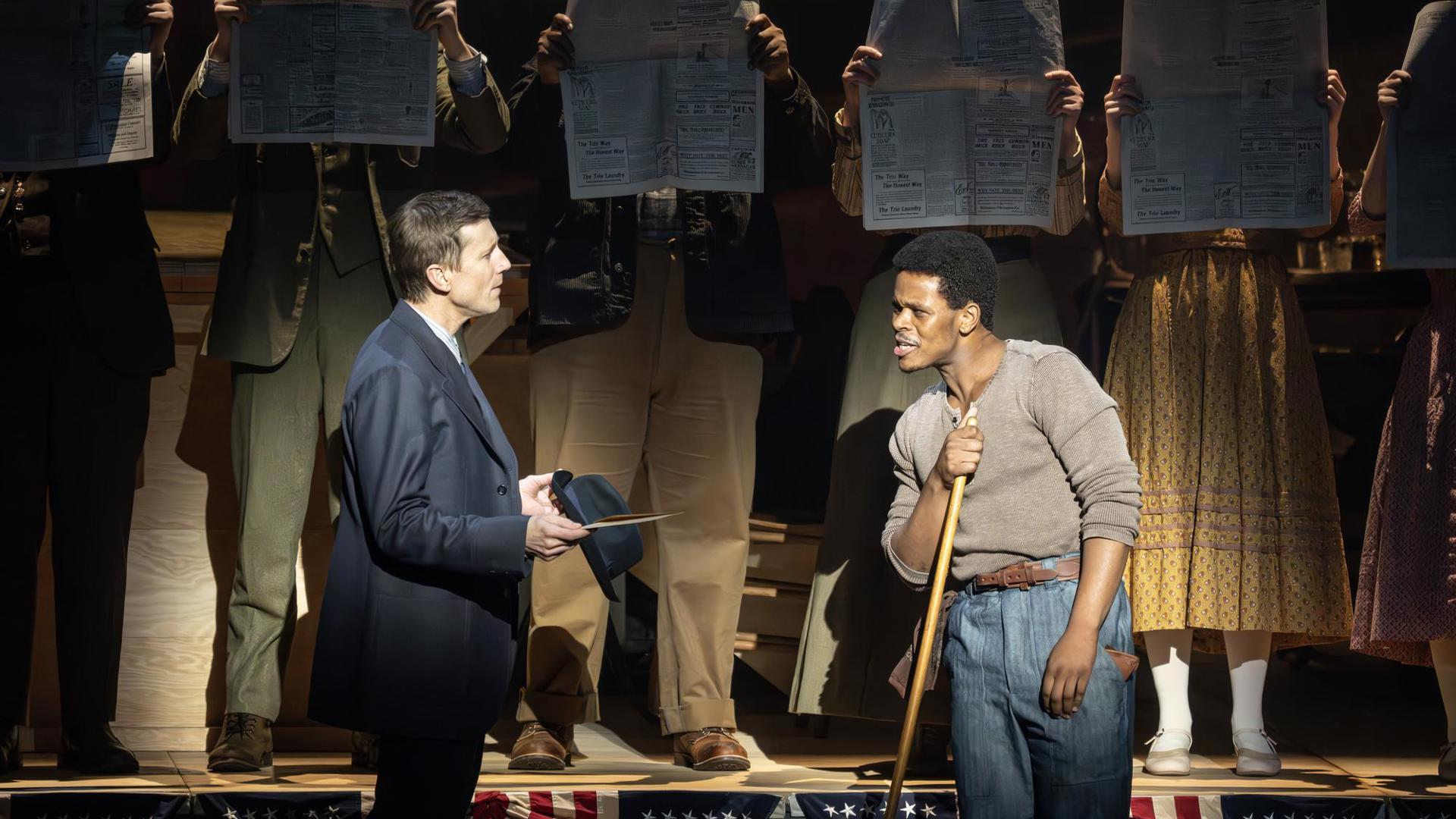 A man with a hat speaks to a man with a broom. Behind them is a line of people with newspapers blocking their faces.