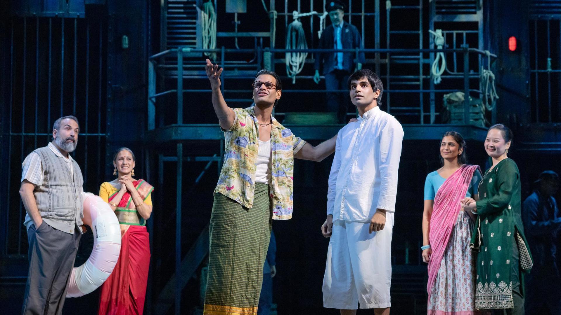 Center stage, an actor gestures out to the audience. Another actor looks to where they are gesturing. In the background, four ensemble members watch the two actors.