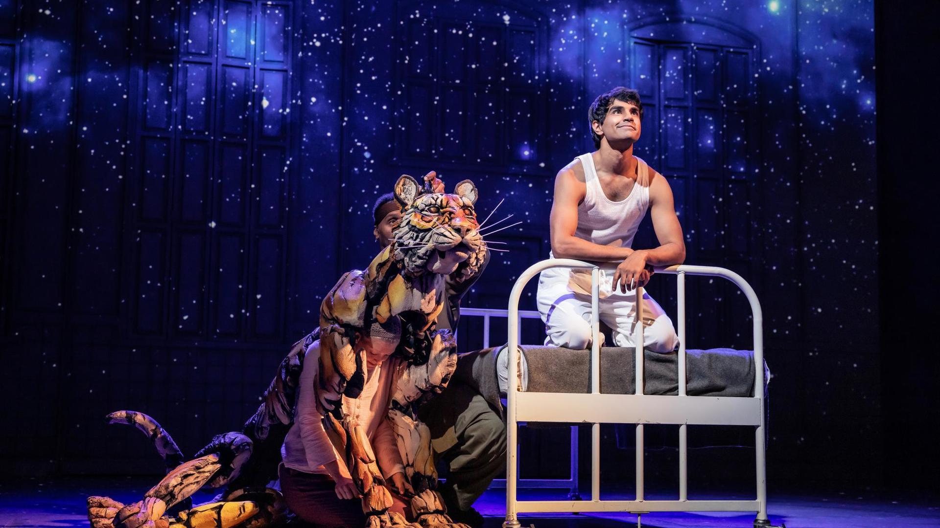 An actor stands on their knees at the edge of a bed set piece. A puppeteer operating a tiger puppet sits on the floor next to them. There are projections of stars on the back wall of the stage.