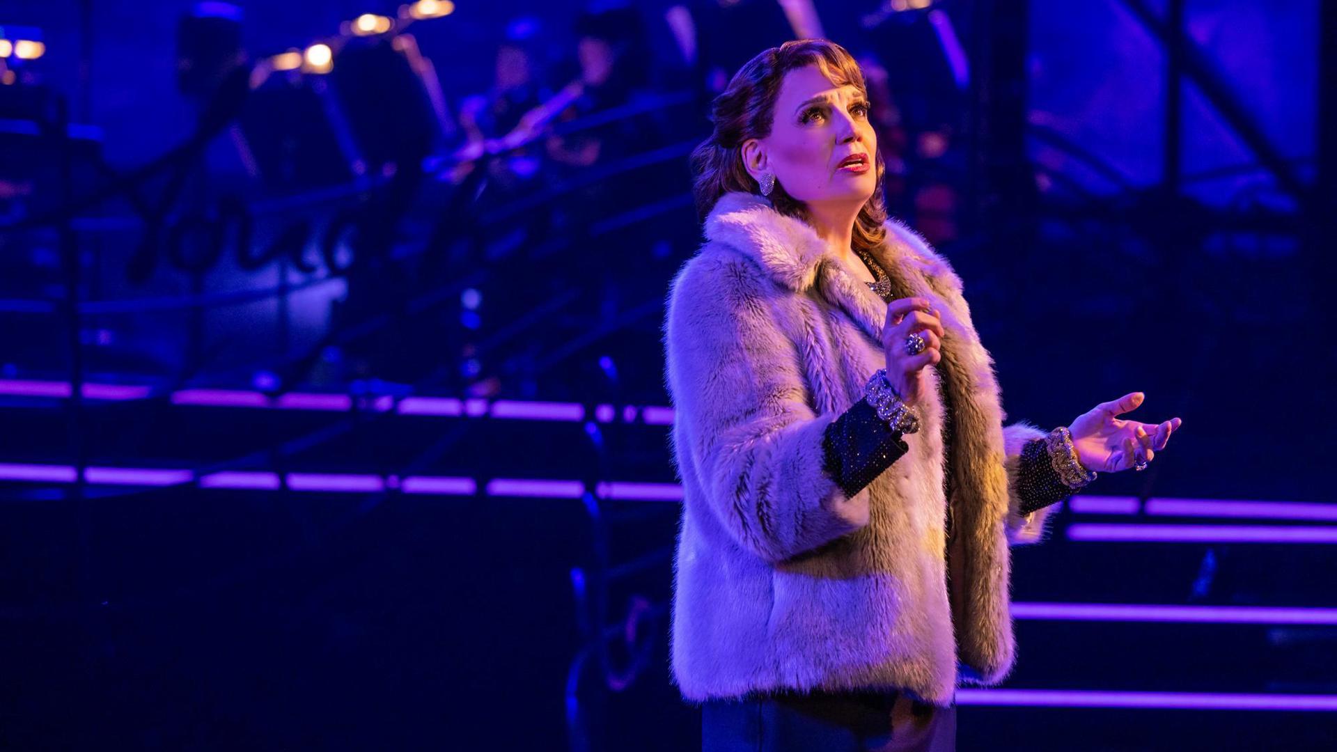 A performer wears a fur coat and lavish jewelry, singing out toward the audience.