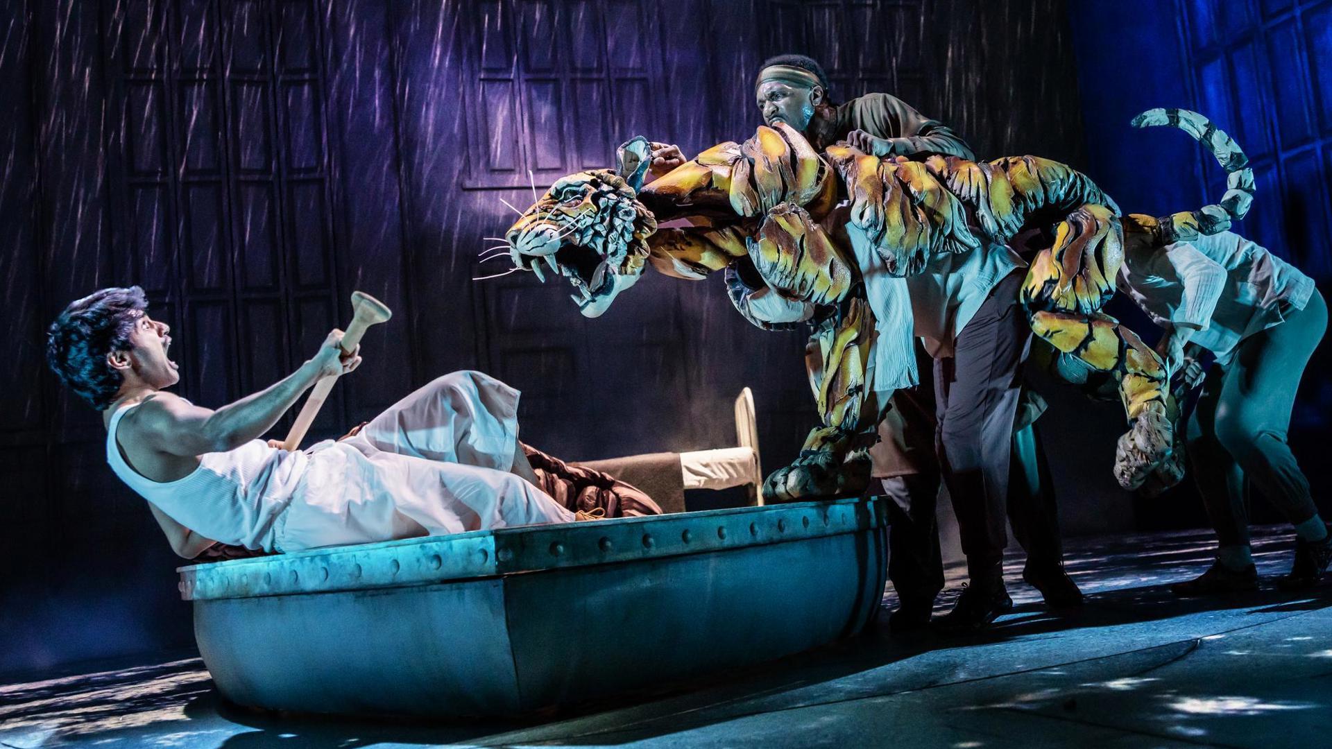 A puppeteer operates a tiger puppet that lunges at an actor. The actor leans backward in a lifeboat holding an oar in defense.