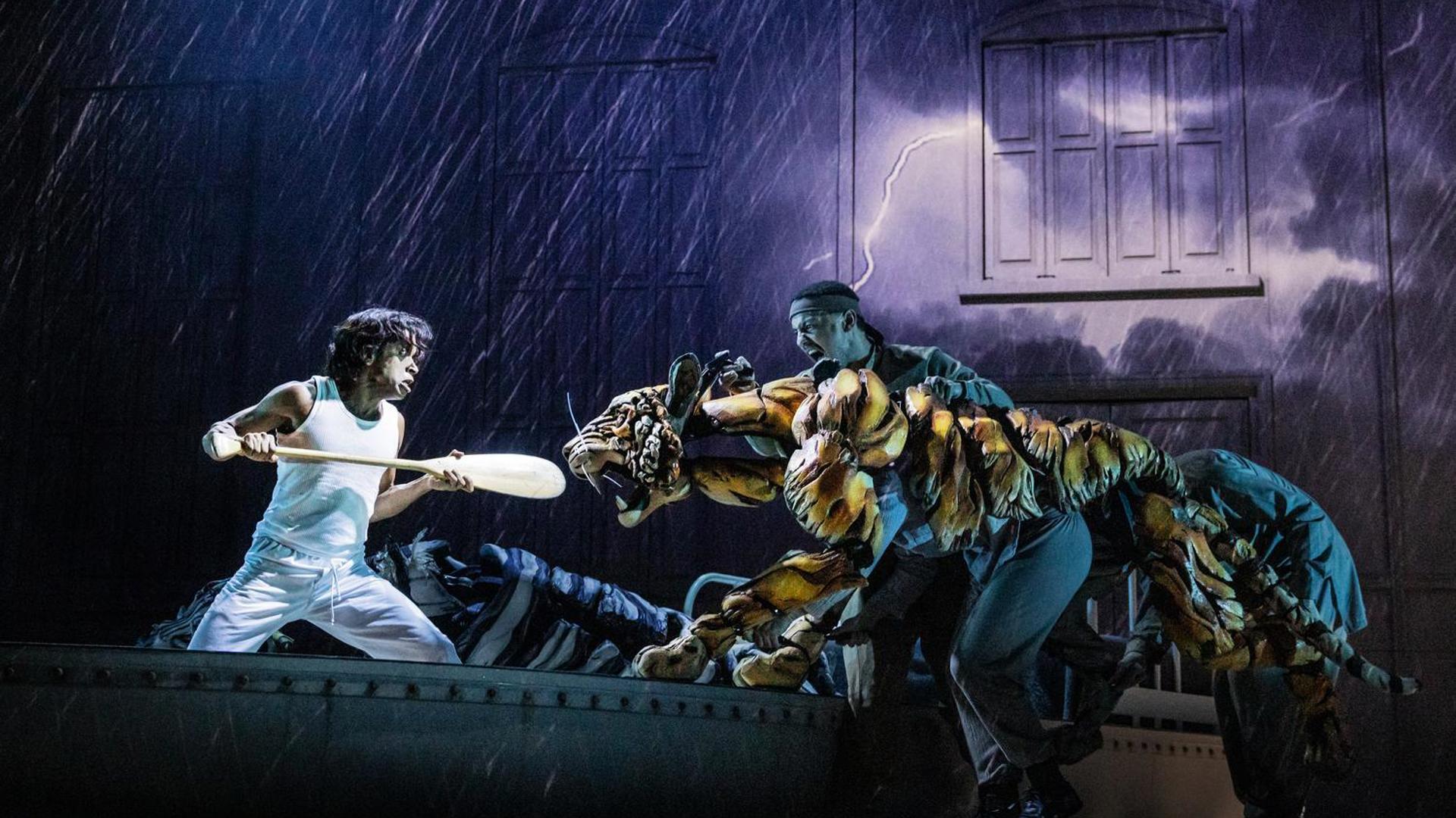 A theatrical scene depicts a person in white using a paddle to fight back a puppet tiger operated by two people, with a stormy backdrop..