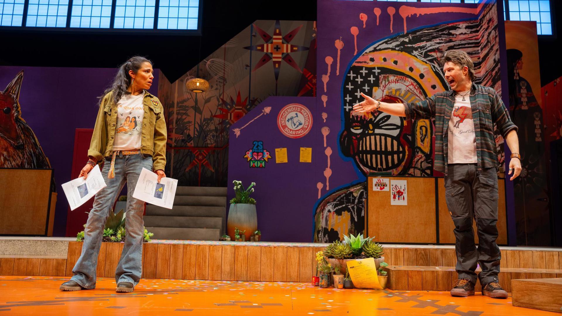 Two people stand on a colorful stage adorned with vibrant Indigenous-inspired artwork, one gesturing passionately while the other holds papers and looks on with concern.