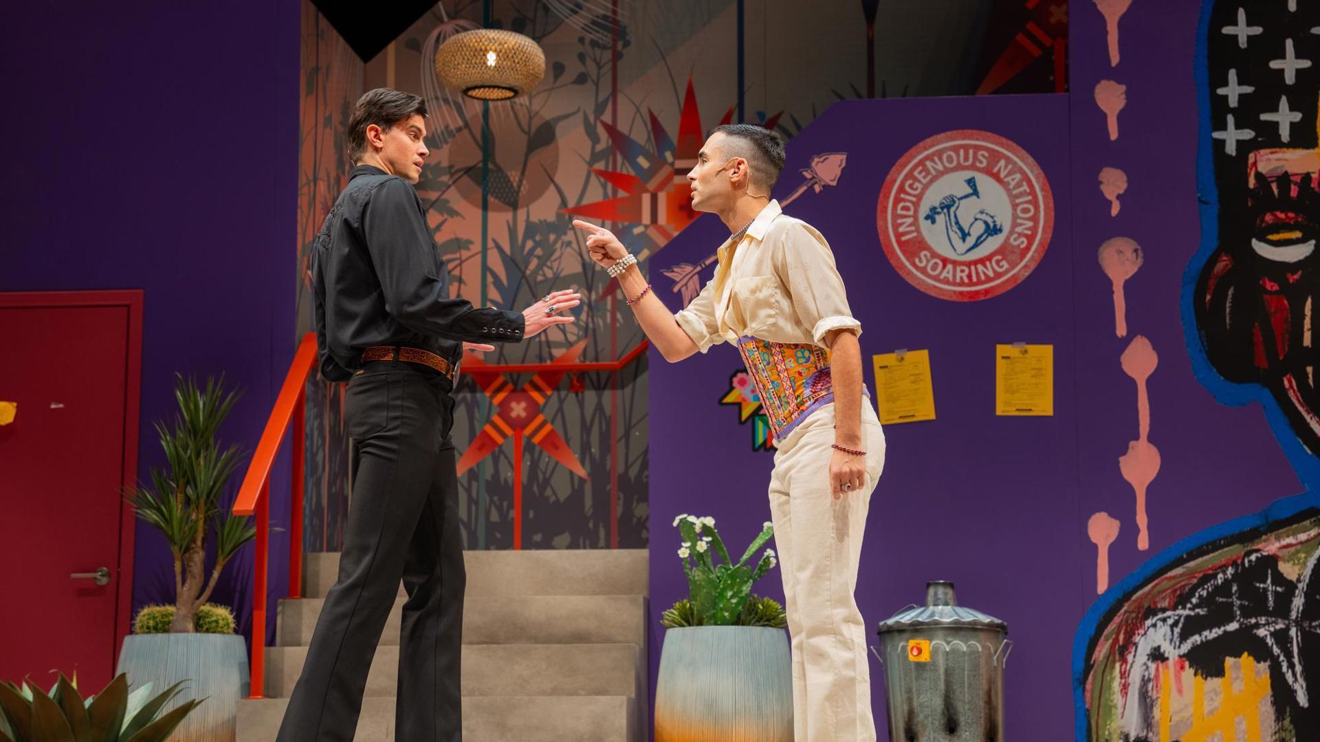 Two people stand on a colorful stage adorned with vibrant Indigenous-inspired artwork, one pointing passionately while the other holds their hand up.